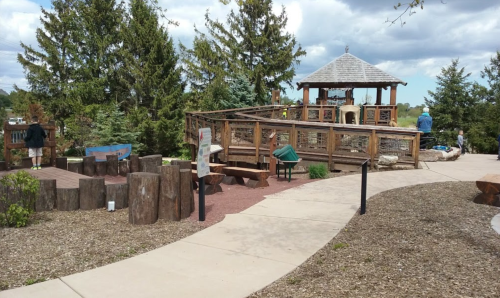 A playground with wooden structures, a gazebo, and benches surrounded by trees and a walking path.