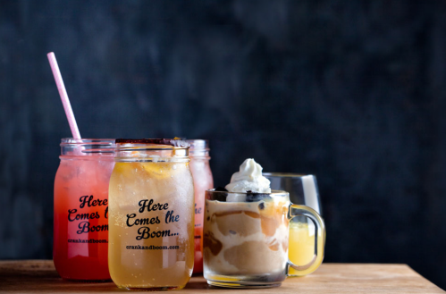 Four drinks on a wooden surface: two colorful cocktails, a clear beverage, and a dessert drink topped with whipped cream.