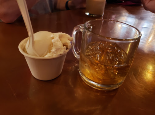 A cup of vanilla ice cream next to a glass of amber-colored whiskey on a wooden table.