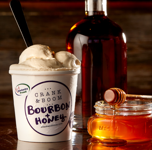 A cup of bourbon and honey ice cream next to a bottle of bourbon and a jar of honey.