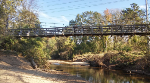 A wooden bridge spans a calm river, surrounded by lush greenery and trees under a clear blue sky.