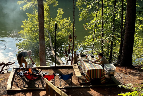A campsite by a lake with a fire pit, a person cooking, and another working at a table under trees.
