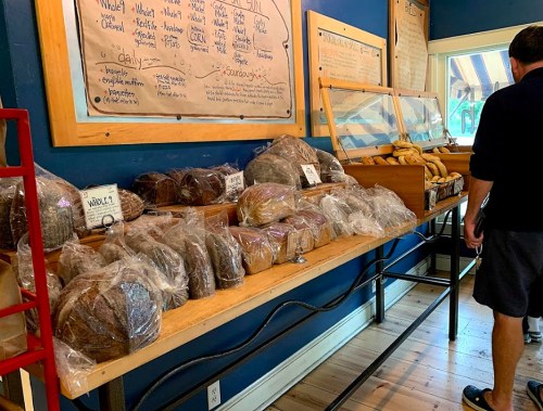 A display of various loaves of bread in bags on a wooden table, with a person browsing in the background.