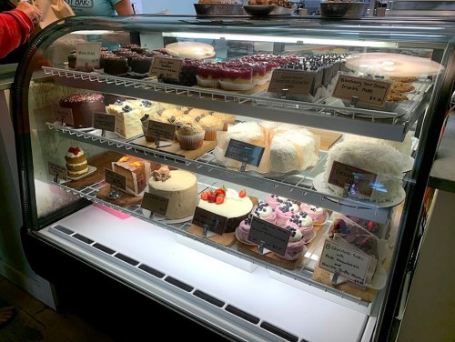 A display case filled with various cakes, cupcakes, and desserts, each labeled with names and prices.