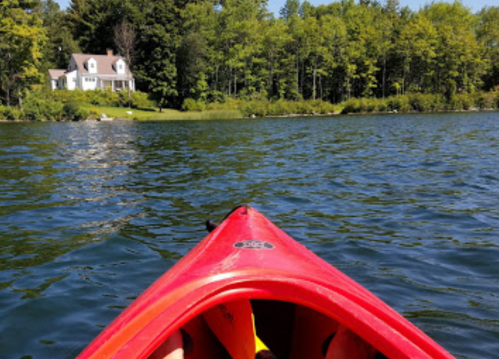 View from a red kayak on a calm lake, with a house and trees in the background.