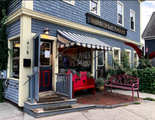 A charming bakery with a blue exterior, red accents, and a striped awning, featuring a red bench outside.