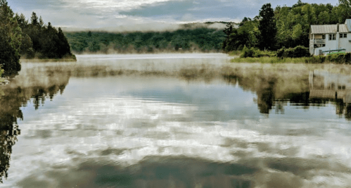 A serene lake reflects misty clouds and trees, with a building visible on the shore.