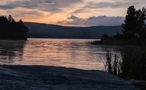 A serene lake at sunset, with calm waters reflecting colorful clouds and surrounded by trees and grass.