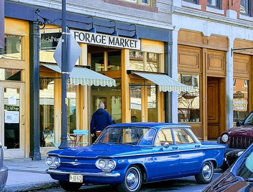 A vintage blue car parked in front of Forage Market, with a person walking by and awnings above the entrance.