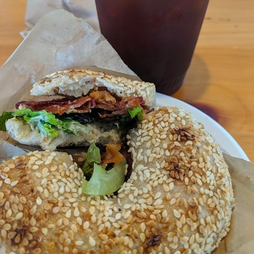 A sesame seed bagel sandwich with lettuce, bacon, and a drink in the background on a wooden table.