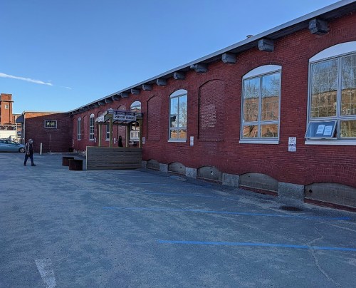 A red brick building with large windows and a wooden entrance, set in a parking area under a clear blue sky.