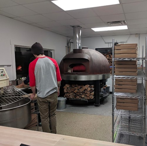 A person in a gray and red shirt stands near a large pizza oven, with stacks of boxes and firewood in the background.