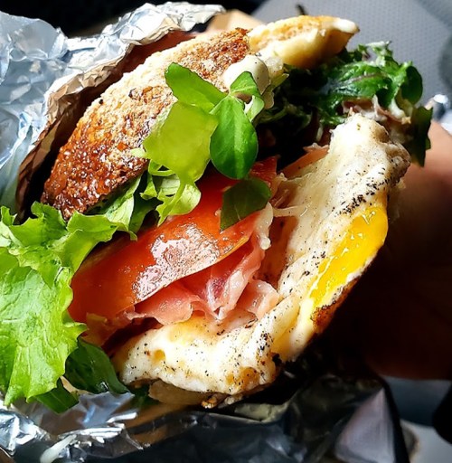 A close-up of a sandwich wrapped in foil, featuring layers of lettuce, tomato, ham, and a fried egg.