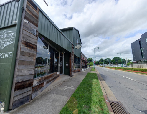 A street view of a rustic building with large windows, greenery, and a cloudy sky in the background.