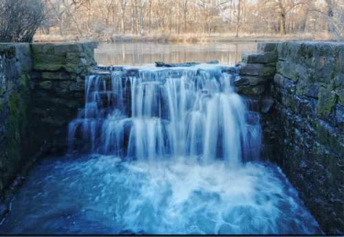 A serene waterfall cascades over stone steps into a calm river, surrounded by trees in a tranquil natural setting.