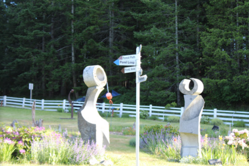 Two abstract sculptures stand near a signpost with directional arrows, surrounded by greenery and flowers.