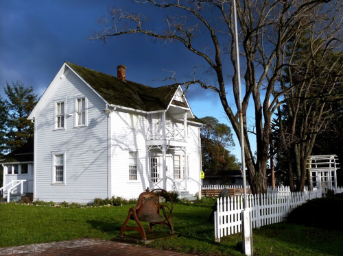 A white two-story house with a porch, surrounded by trees and a picket fence, under a cloudy sky.