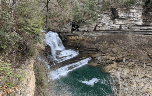 A serene waterfall cascades over rocky cliffs into a tranquil green pool, surrounded by trees and natural rock formations.