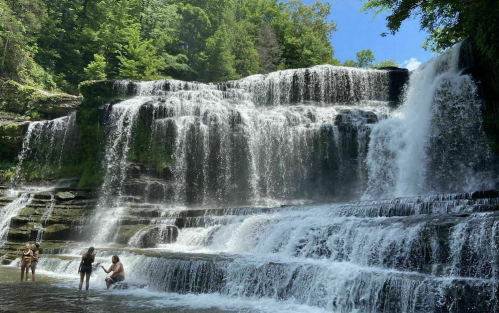 A scenic waterfall with people enjoying the water, surrounded by lush green trees and a clear blue sky.