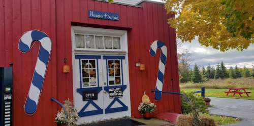 Red school building with candy cane decorations, open sign, and picnic table outside, surrounded by trees and grass.