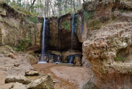 A serene landscape featuring two waterfalls cascading over rocky cliffs into a sandy area surrounded by trees.