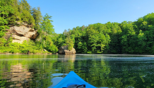 A serene view from a kayak on calm water, surrounded by lush green trees and rocky formations under a clear blue sky.