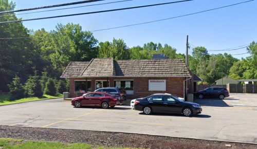 A brick building with a sloped roof, surrounded by trees, featuring parked cars in the lot on a sunny day.
