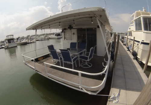 A houseboat with a dining area on the deck, surrounded by other boats in a marina under a cloudy sky.