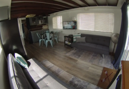 Cozy living room with a gray sofa, blue dining set, and a TV, featuring wooden beams and large windows.