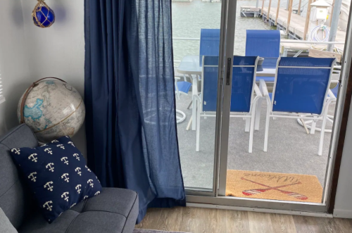 Cozy interior with a gray couch, globe, blue curtains, and a view of blue chairs on a dock through a sliding glass door.