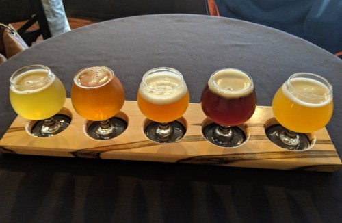 A wooden flight tray holds five different craft beers in small glasses, arranged on a dark table.