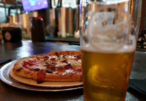 A close-up of a pizza on a plate with a glass of beer in the foreground, set in a bar environment.