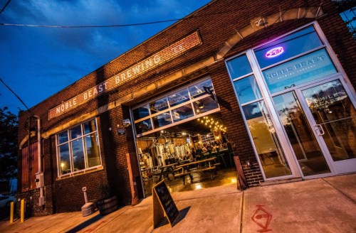 Exterior of Noble Beast Brewing, featuring a brick facade, large windows, and a neon open sign at dusk.