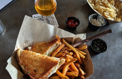 A plate with a grilled cheese sandwich, fries, and dipping sauces, alongside a glass of beer and a bowl of chips.