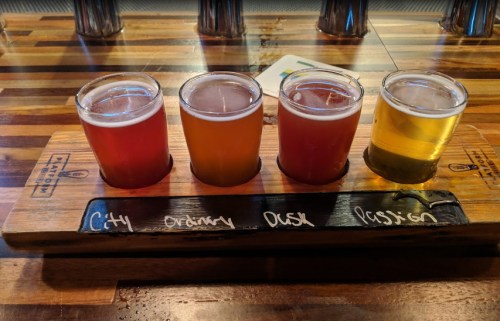 A wooden flight board with four small glasses of different colored beers labeled "City," "Ornery," "Dusk," and "Passion."