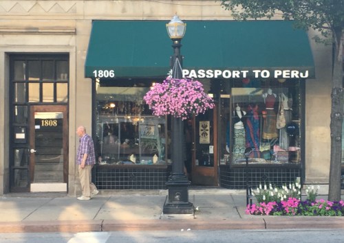 A man walks past a shop called "Passport to Peru," adorned with colorful flowers and a green awning.