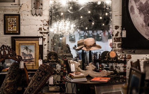 A vintage shop interior featuring a mirror, a hat, and various antique items displayed on tables and walls.