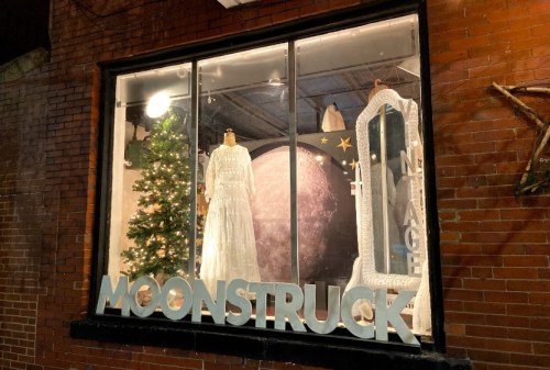 A storefront window display featuring a white dress, a Christmas tree, and a large moon backdrop with "MOONSTRUCK" sign.