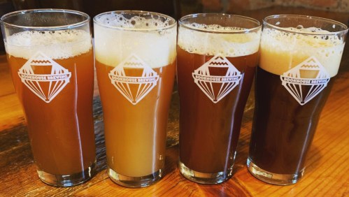 Four glasses of craft beer with varying colors and foam, displayed on a wooden table.