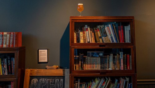 A glass of beer sits atop a wooden bookshelf filled with books, next to a chalkboard sign that says "OPEN."
