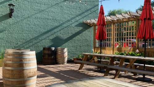 Outdoor patio with wooden barrels, picnic tables, and red umbrellas against a green brick wall.