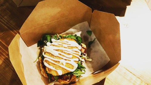 A sandwich in a takeout box, layered with greens, a crispy patty, and drizzled with white sauce.
