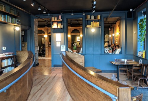 Cozy café interior with wooden furniture, bookshelves, and warm lighting, featuring a view of patrons in a back room.