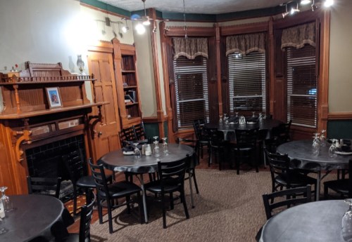 Cozy restaurant interior with dark tables, wooden accents, and large windows, featuring a fireplace and decorative shelves.