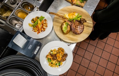 A kitchen counter displaying gourmet dishes: grilled meats, vegetables, and a side of pasta, with a receipt printer nearby.