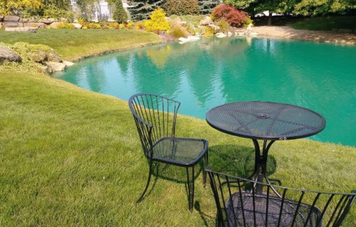 A black metal table and chair set on green grass by a tranquil turquoise pond surrounded by colorful plants.