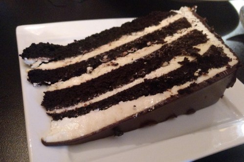 A slice of layered chocolate cake with alternating layers of dark chocolate and creamy frosting on a white plate.