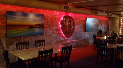 Interior of a restaurant featuring stone walls, colorful artwork, and dim lighting with tables and chairs arranged.