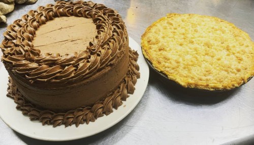 A chocolate frosted cake with decorative icing next to a golden brown pie on a silver countertop.