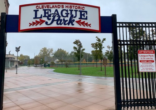 Entrance to Cleveland's Historic League Park, featuring a sign and a view of the green field beyond, on a rainy day.
